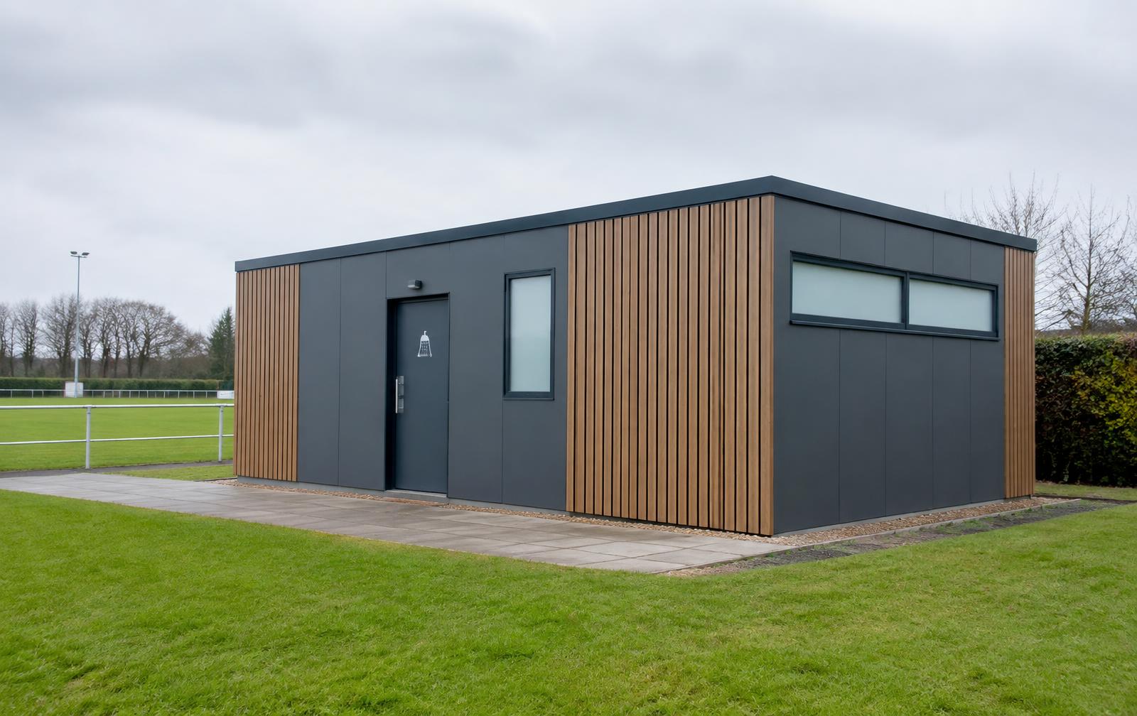 Modular shower building with grey panels and timber-effect cladding beside a sports pitch